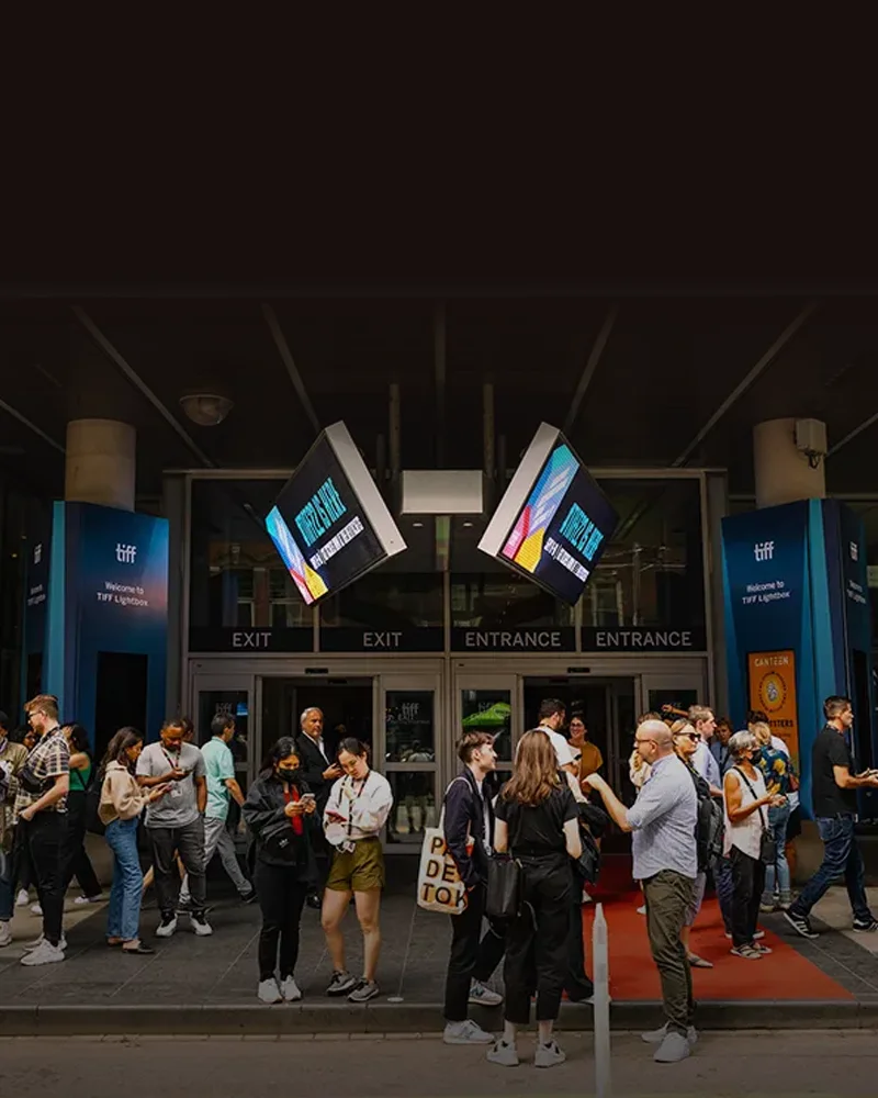 Crowd of people gathered outside TIFF Lightbox entrance with glowing digital signs overhead.
