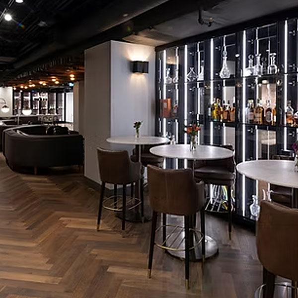 Modern bar interior with illuminated liquor display, high tables, brown leather stools and herringbone wood floor