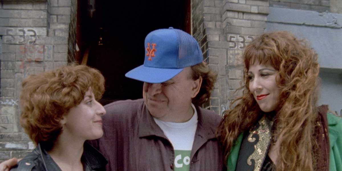 Three people pose together outside a brick building, man in blue NY Mets cap flanked by two women with curly red hair.