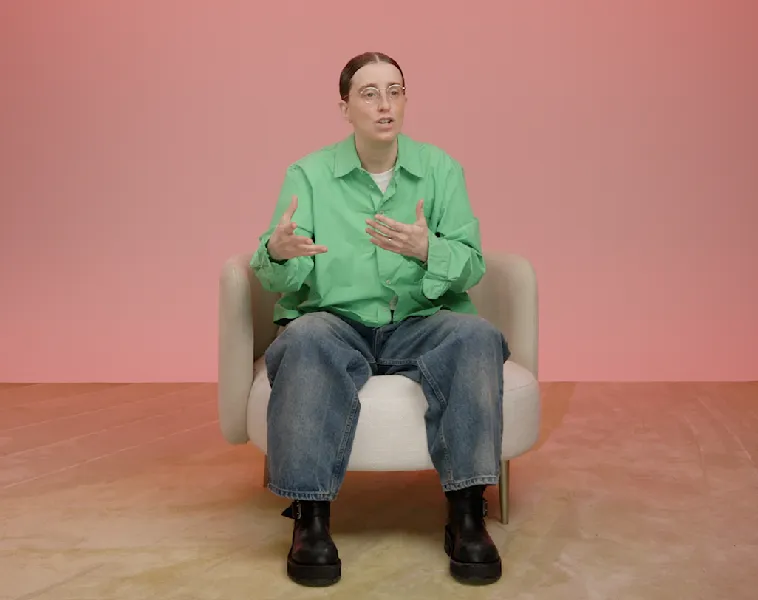 A person in a green shirt and jeans sits in a white chair, gesturing while speaking against a pink background.