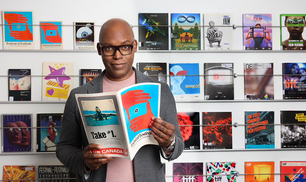 Cameron Bailey, CEO of TIFF holding the first Festival programming book and standing in front of a wall covered with various Festival programme books over the years