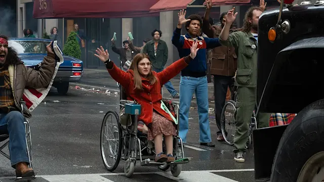 Woman in red coat in wheelchair raises arms during a street protest, surrounded by a crowd.