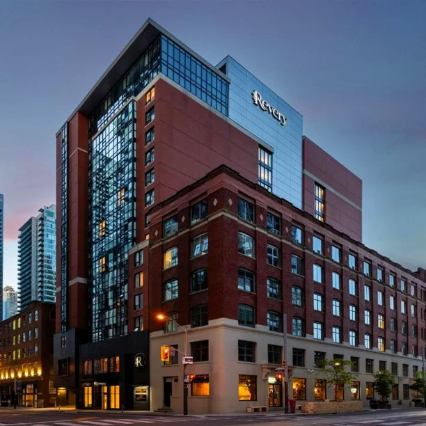 Modern hotel building with red brick base and glass tower addition, illuminated at dusk in urban setting
