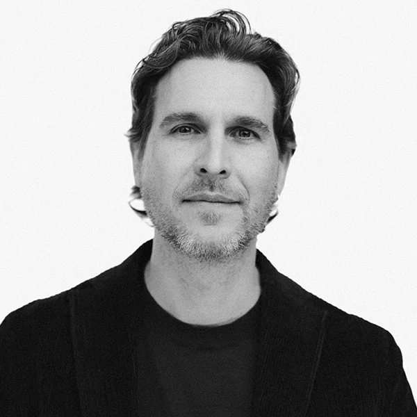 Black and white headshot of a man with short wavy hair, stubble, wearing a dark blazer.