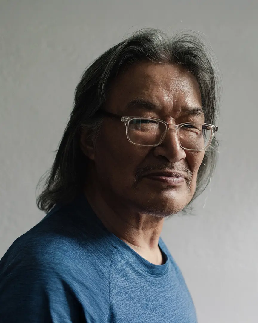 Close-up portrait of a person with long dark hair and glasses wearing a blue shirt against a light background