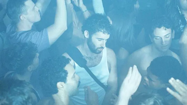 A crowd of people dancing in a blue-lit nightclub, with a bearded man in a white tank top at center.