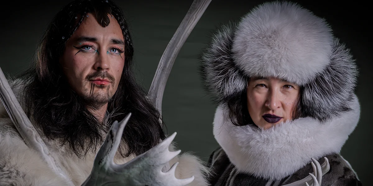 Two people in traditional fur clothing and dramatic makeup pose with antlers against a dark background.