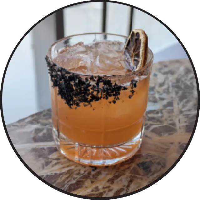 Orange cocktail in rocks glass with black salt rim and cinnamon stick garnish on marble surface