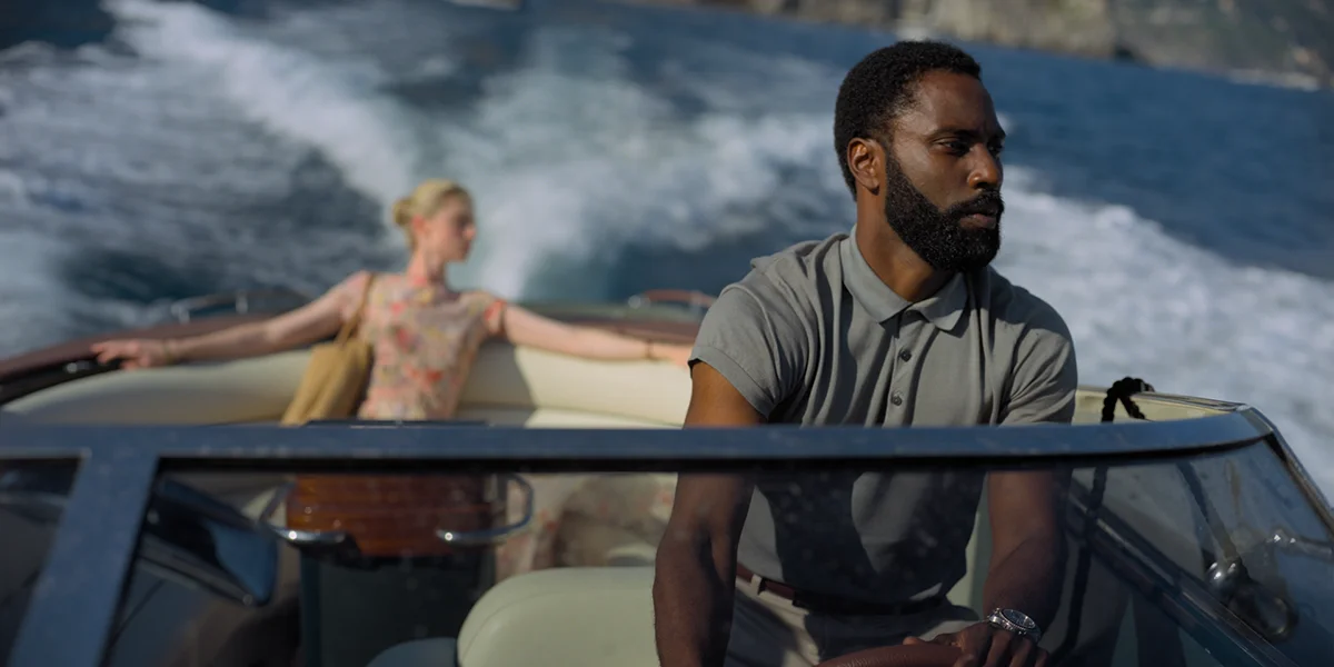 A man in a gray polo sits at the front of a speedboat while a woman stands with arms outstretched behind him.