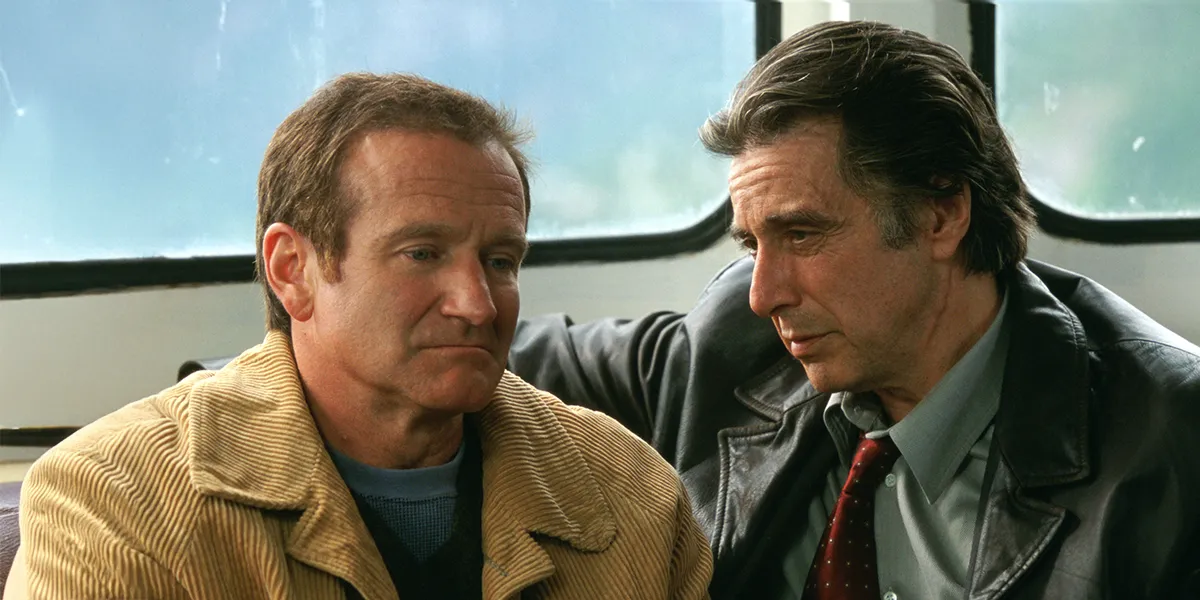 Two men sit together in a tense conversation, one in a tan jacket, the other in a dark leather jacket and red tie.