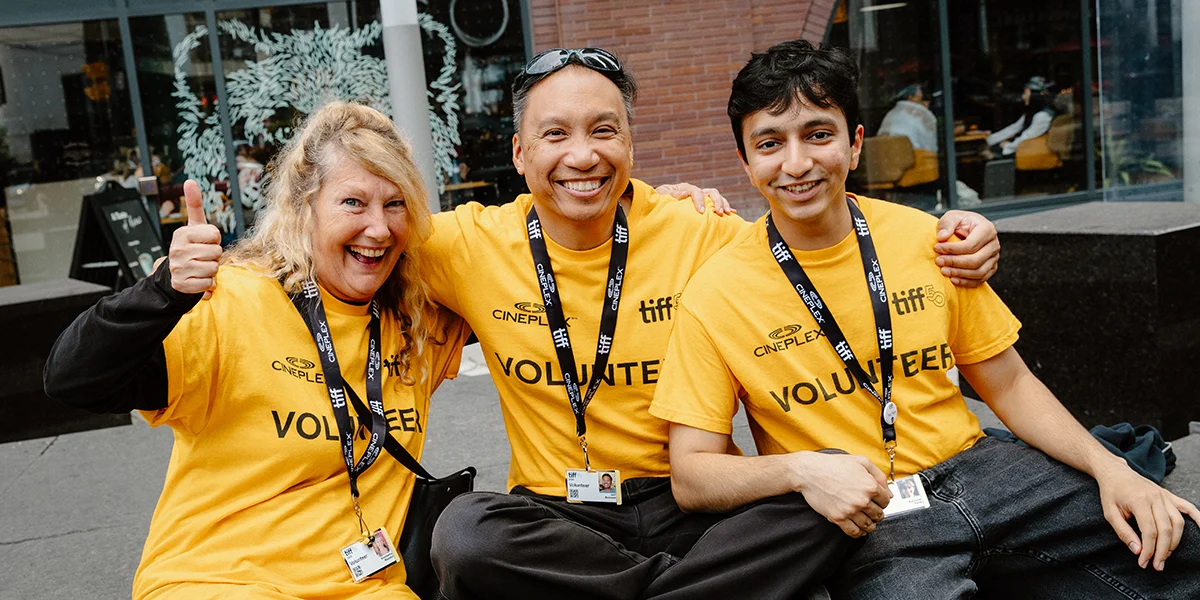 Three smiling TIFF volunteers in yellow Cineplex shirts pose together outdoors, one giving a thumbs up.