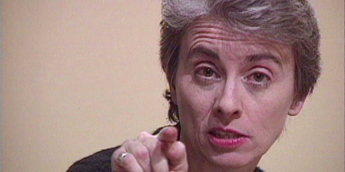 A middle-aged woman with short gray hair points her finger forward, speaking expressively against a beige background.