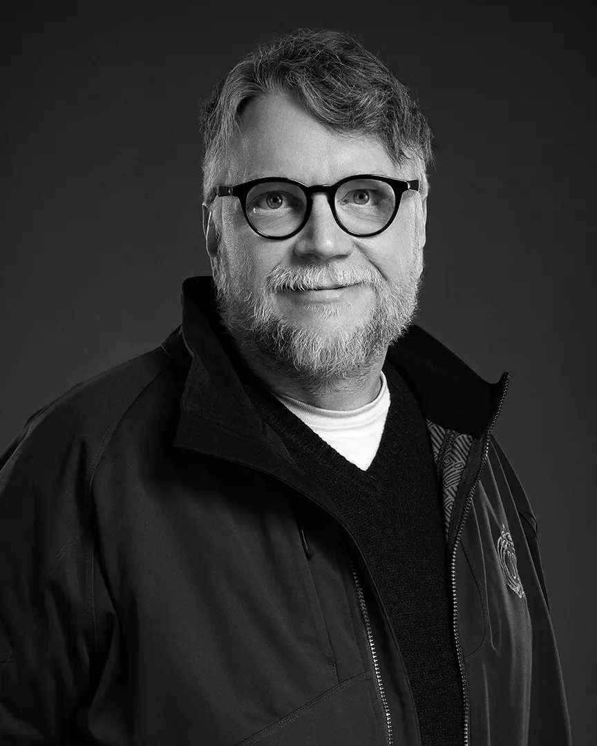 Black and white portrait of Guillermo del Toro wearing glasses and a dark jacket, smiling warmly at the camera