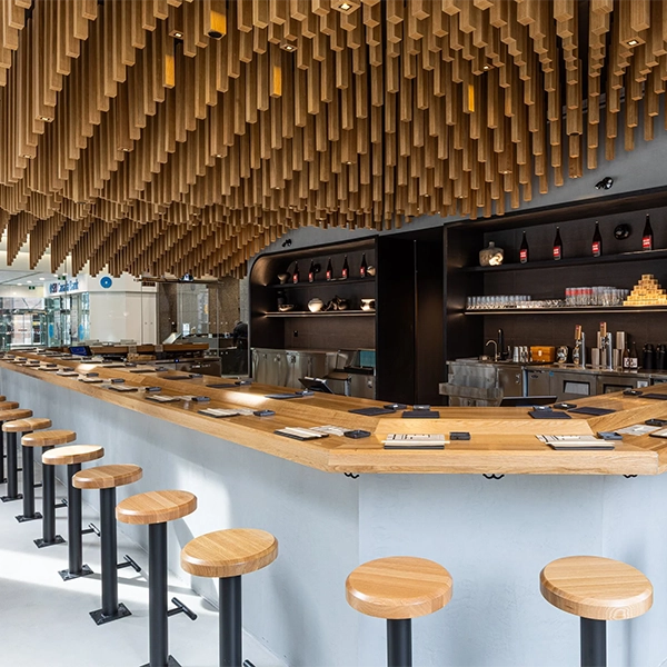 Modern sushi bar featuring a wraparound wooden counter lined with backless stools underneath an ornate ceiling installation of hanging wooden beams.