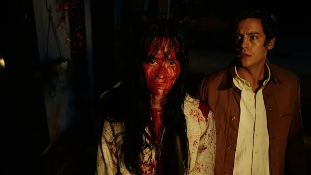 A woman covered in blood stands beside a frightened man in a dark, eerie horror film scene.