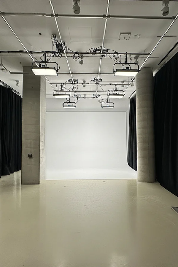 Empty photography studio with white backdrop, overhead lights, black curtains, and concrete columns.