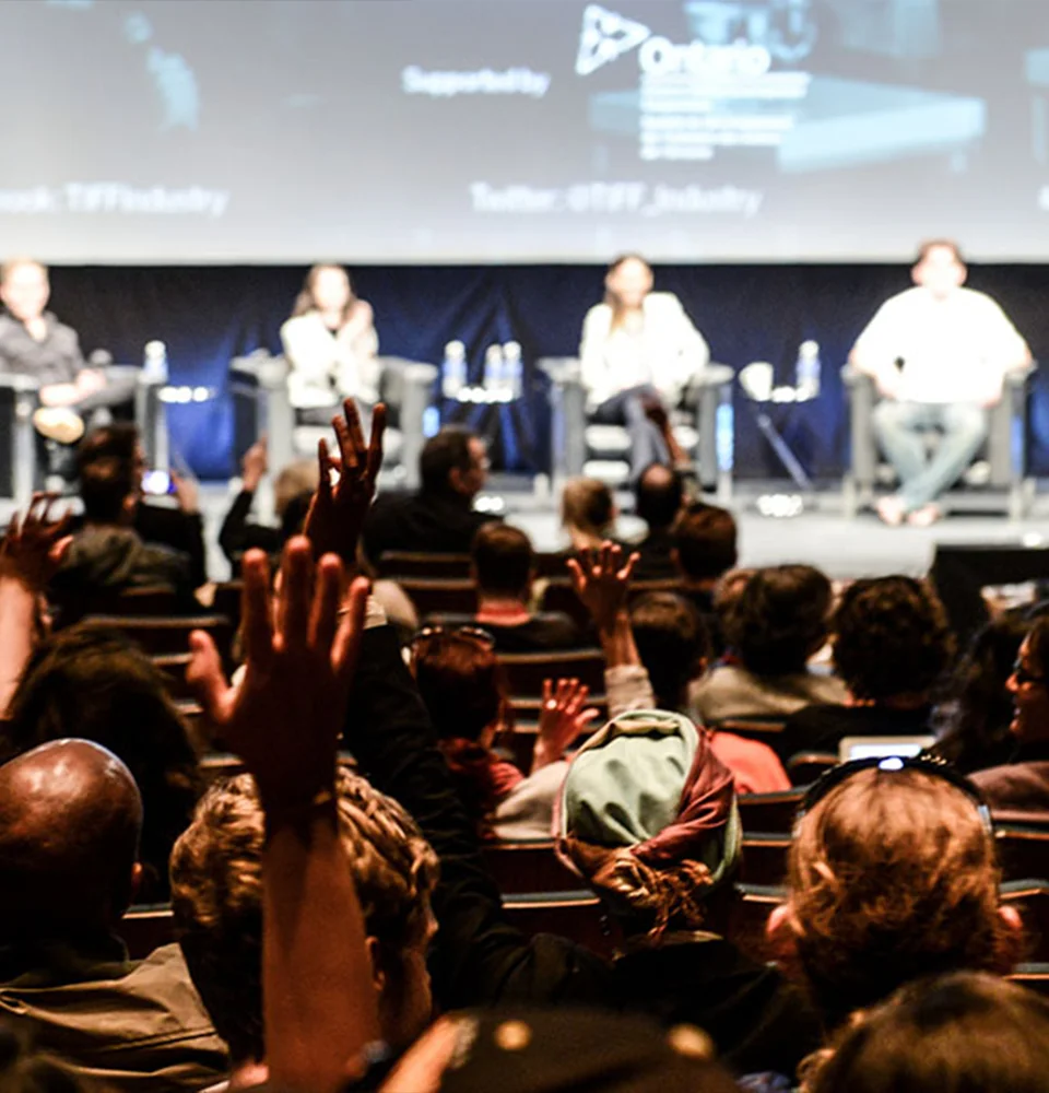 Panel discussion on stage with audience members raising hands during Q&A session in darkened auditorium