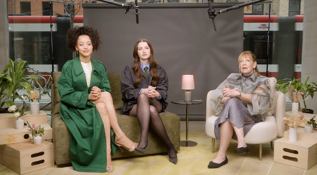 Three women are seated in a studio setting with plants, microphones, and a small lamp nearby.