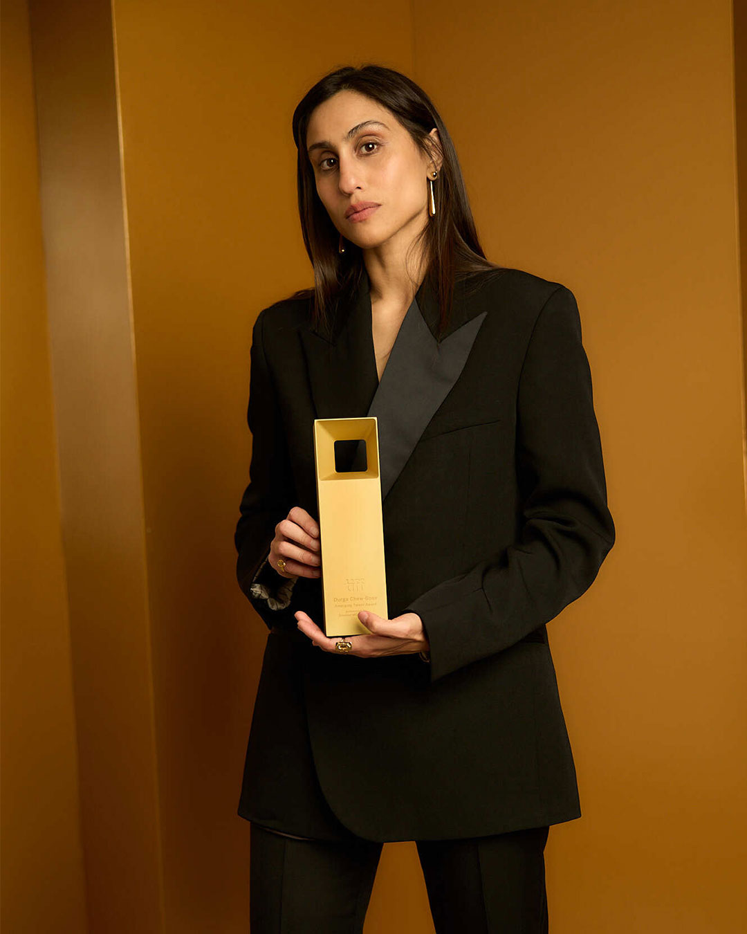 Portrait of Durga Chew-Bose in a black suit standing and holding the award