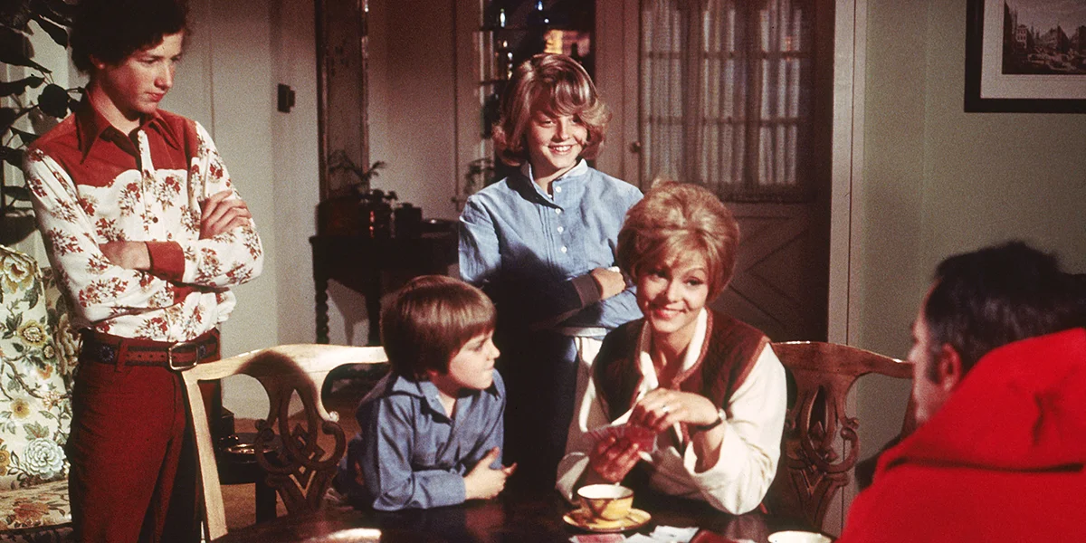A woman and children gather around a table in a warm, vintage 1970s living room scene.