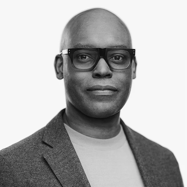 Black and white portrait of a bald man wearing bold-framed glasses and a blazer, smiling slightly.