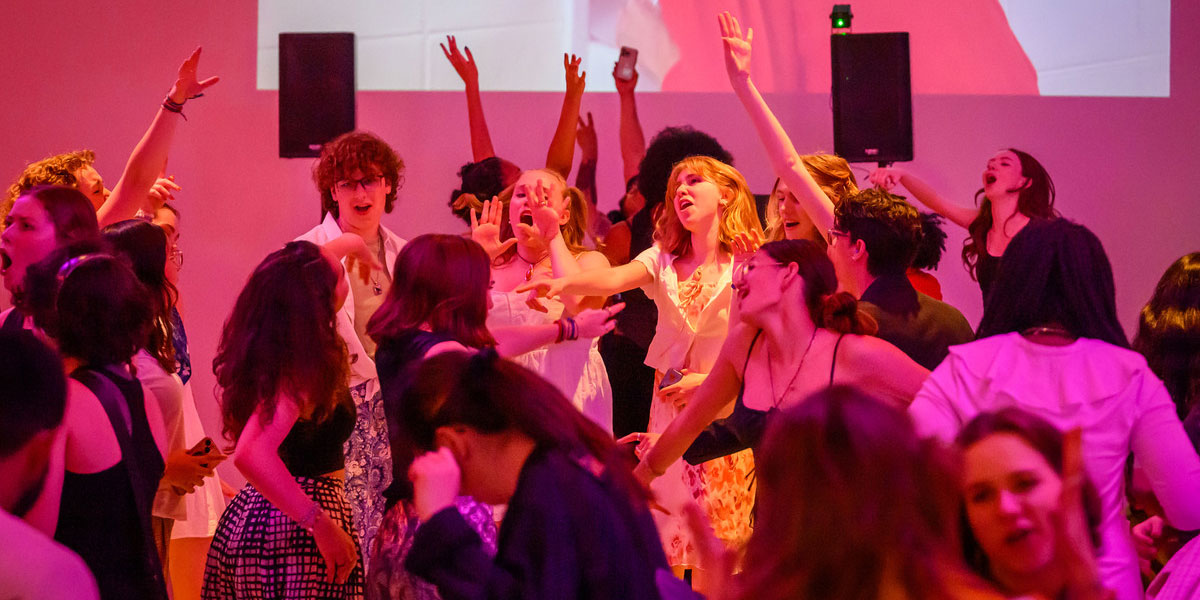 A bunch of teens dancing and rocking out in an auditorium bathe in pink light