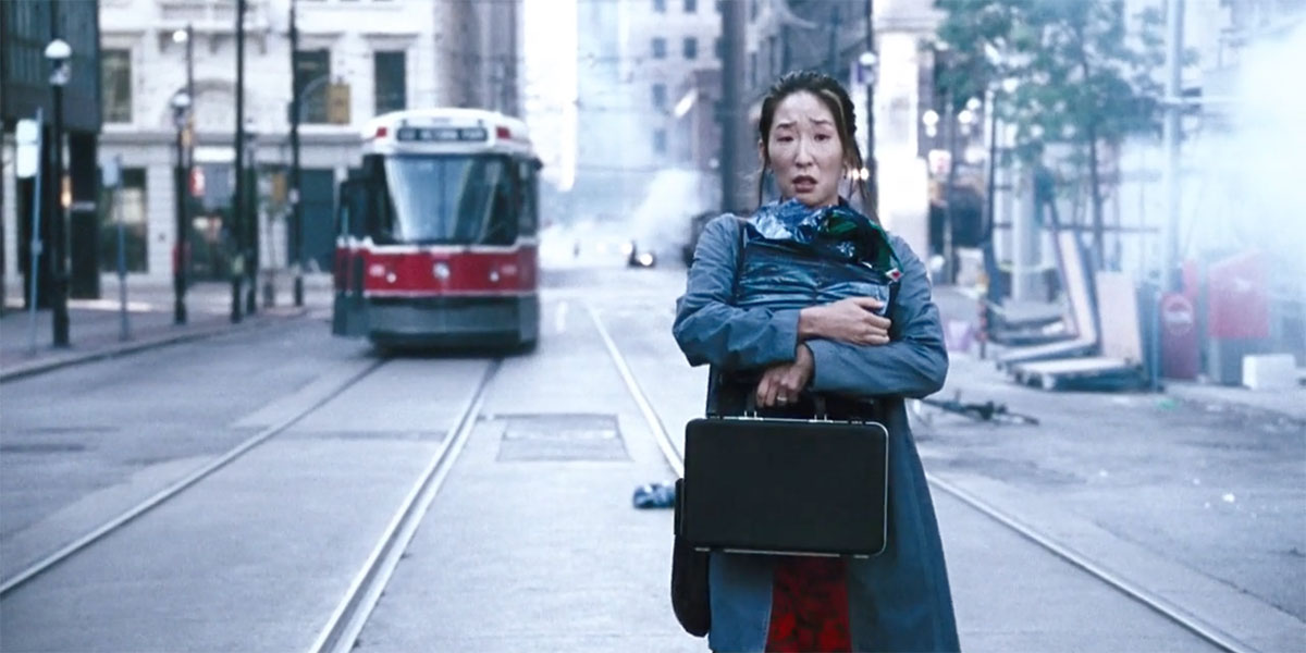 A woman hugging a bag and holding onto a briefcase walks down a street as a streetcar appears behind her