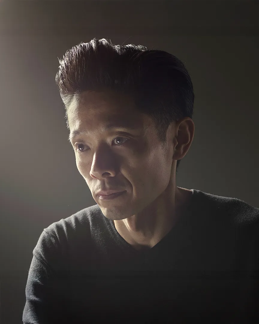 Dramatic side-lit portrait of a person in a black sweater against a dark background, with a contemplative expression