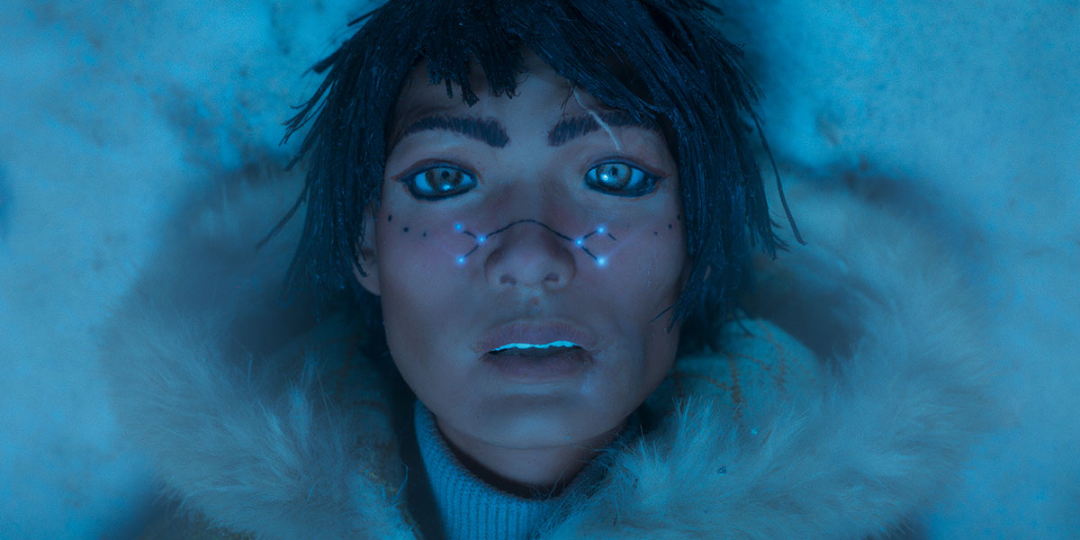 A Indigenous women in a parka lying on the snow covered ground looking up to the camera