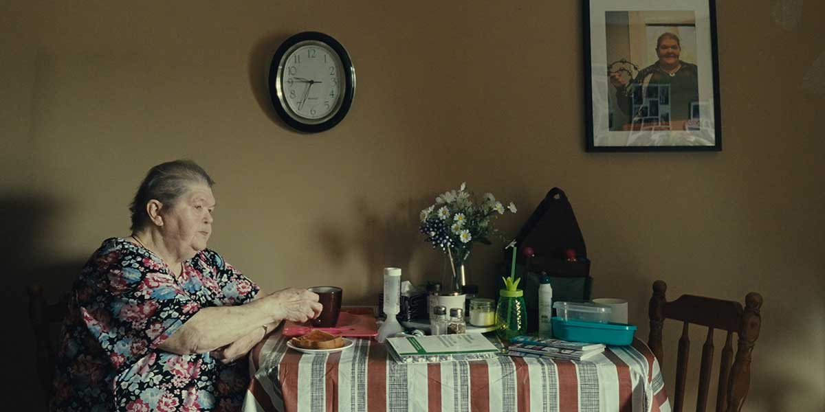 Elderly woman in floral dress sits at striped tablecloth with coffee, flowers, and medications on table