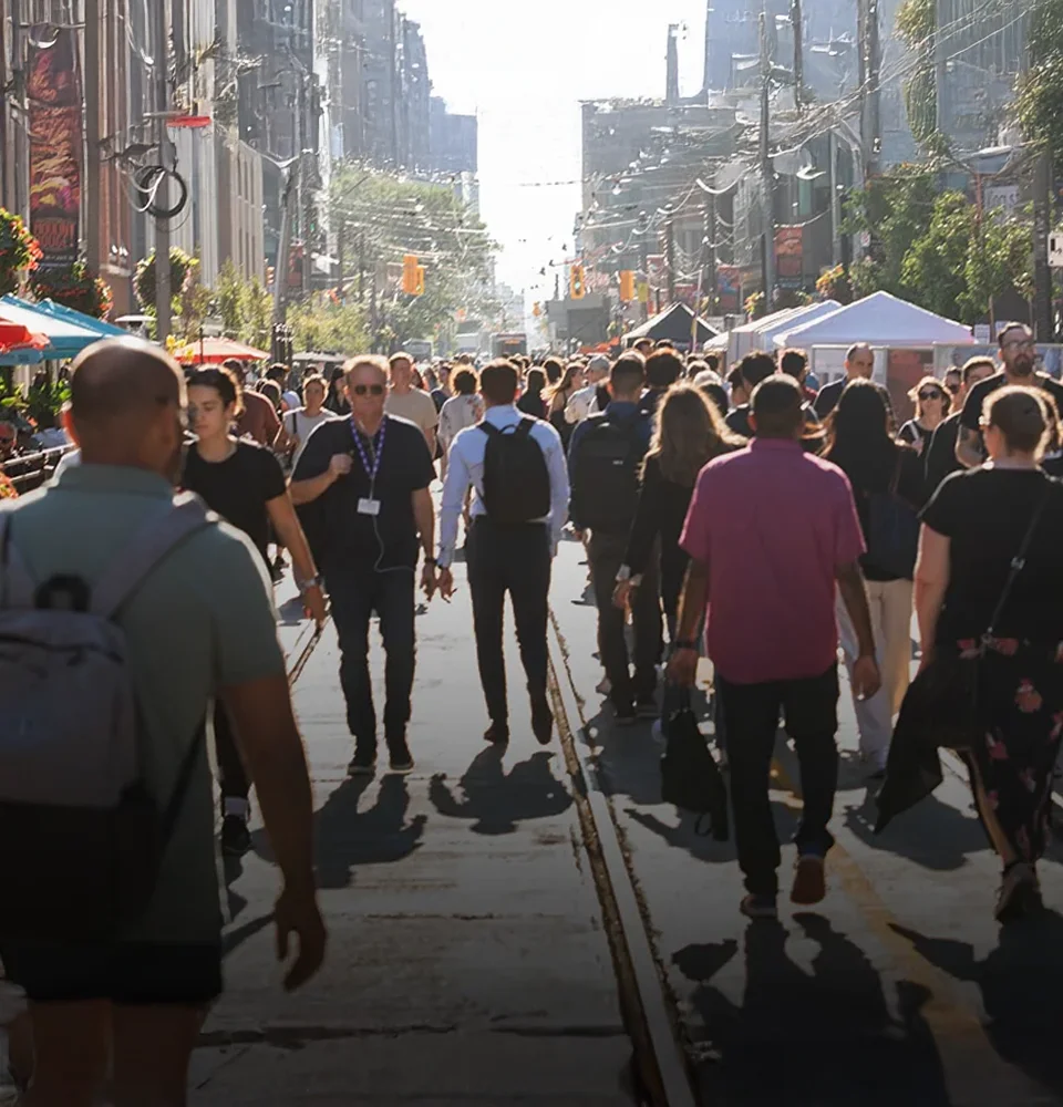 A large crowd of people walking along a busy city street on a sunny day with vendor tents.