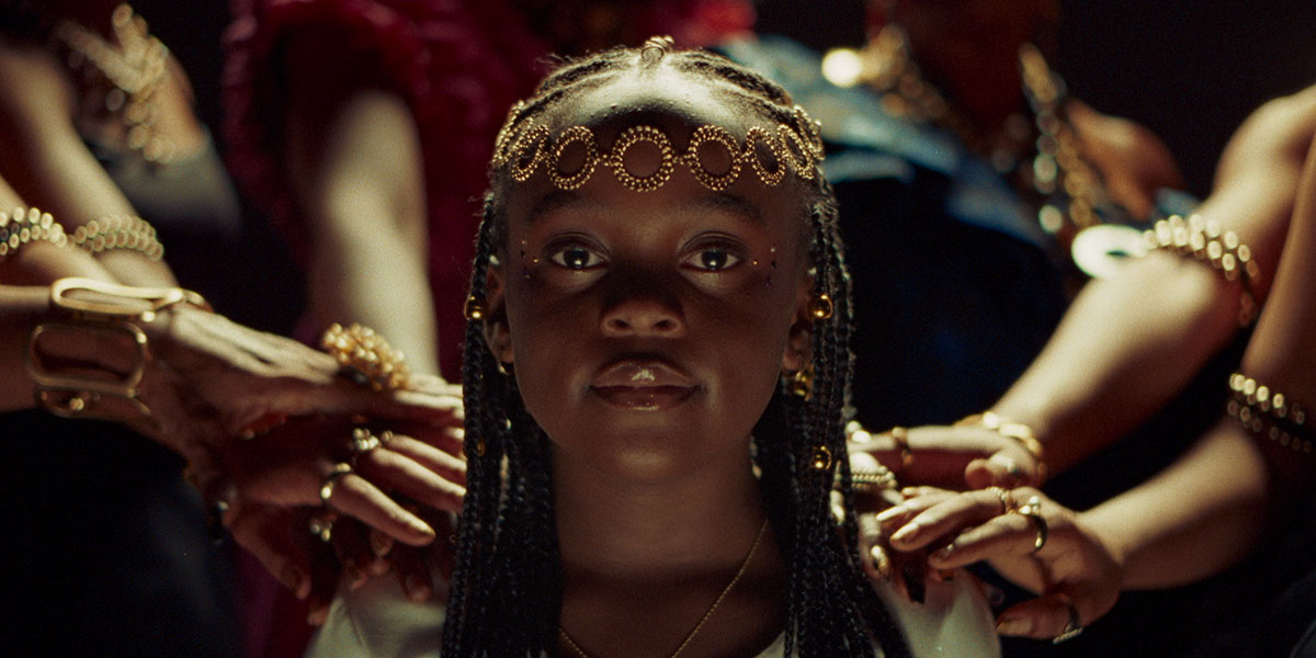 A young Black ballerina in costume looking at the camera with several hands touching her shoulders