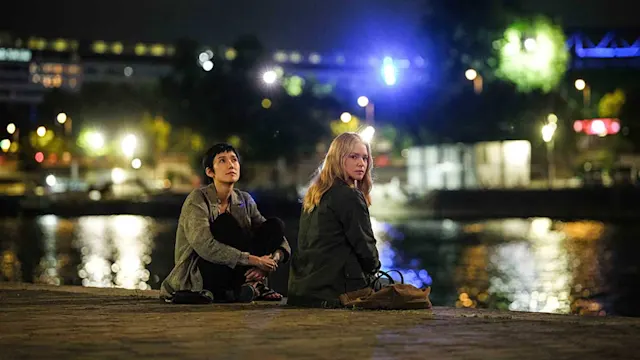 Two people sit by a river at night, city lights reflecting on the water behind them.