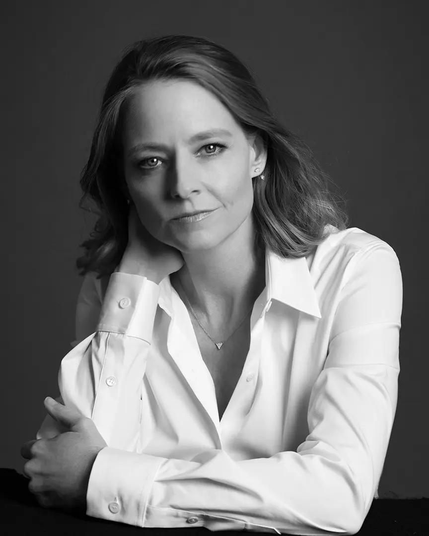 Black and white portrait of Jodie Foster in a white button-down shirt with a subtle smile against a dark background