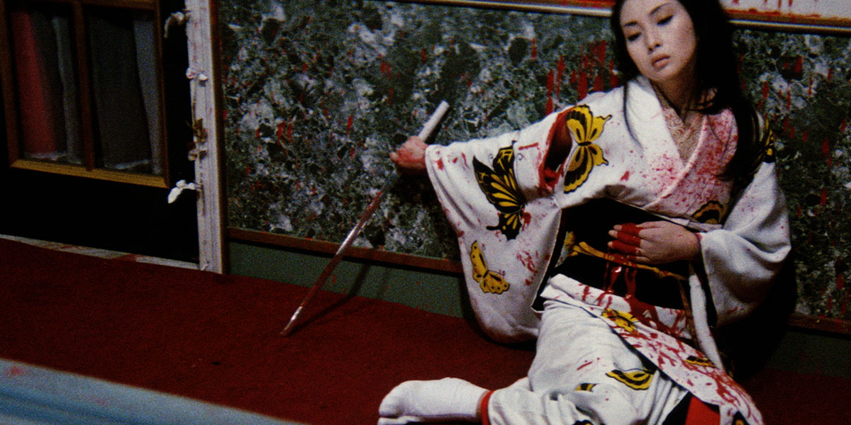 Yuki Kashima (Meiko Kaji) on the floor against the wall holding her hand on her stomach to try and stop the bleeding