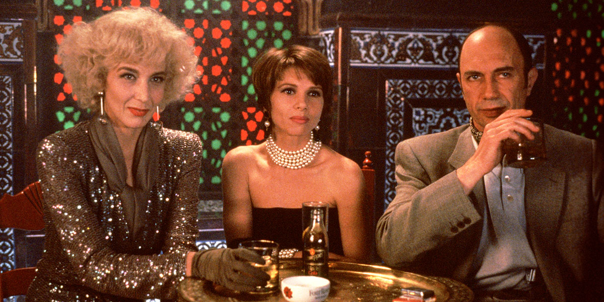 Two women, one in a sparkling dress and the other in a black strapless dress are sitting with  a man in a grey suit inside a bar