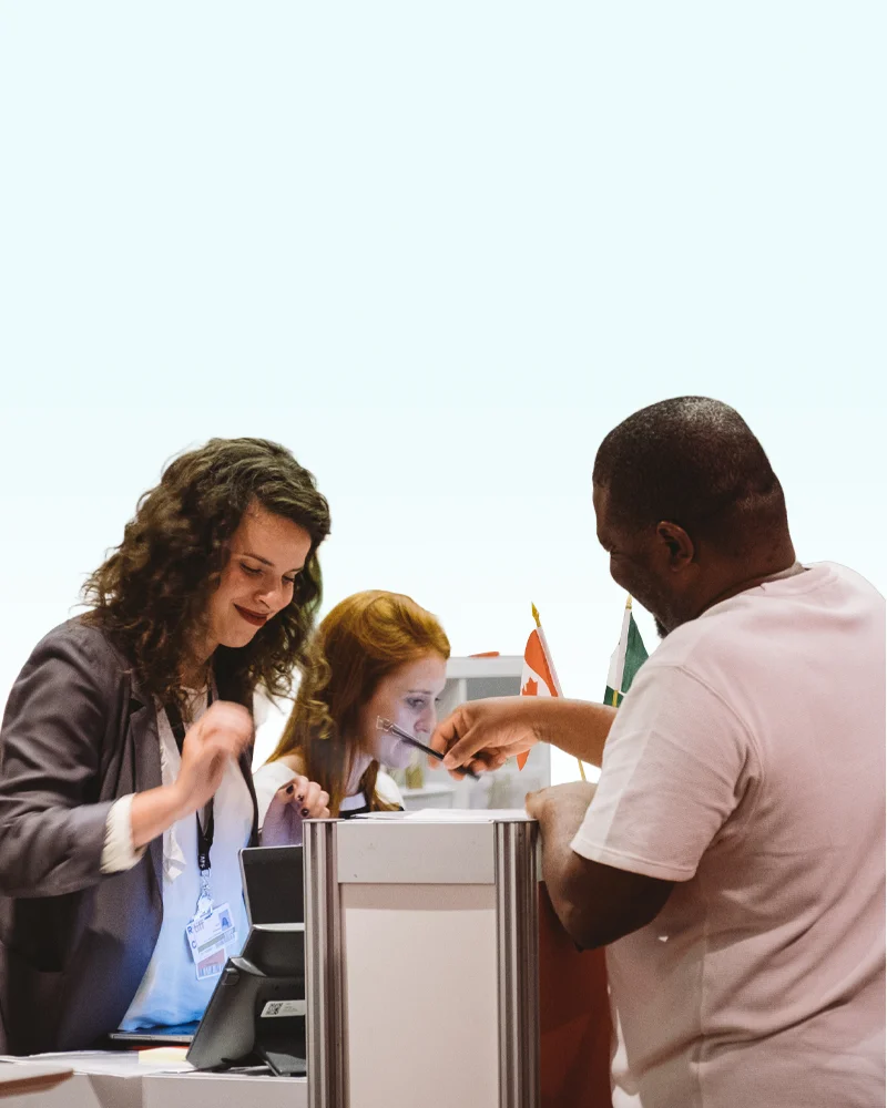 Two women at a registration desk assist a man at an event with small country flags in the background.
