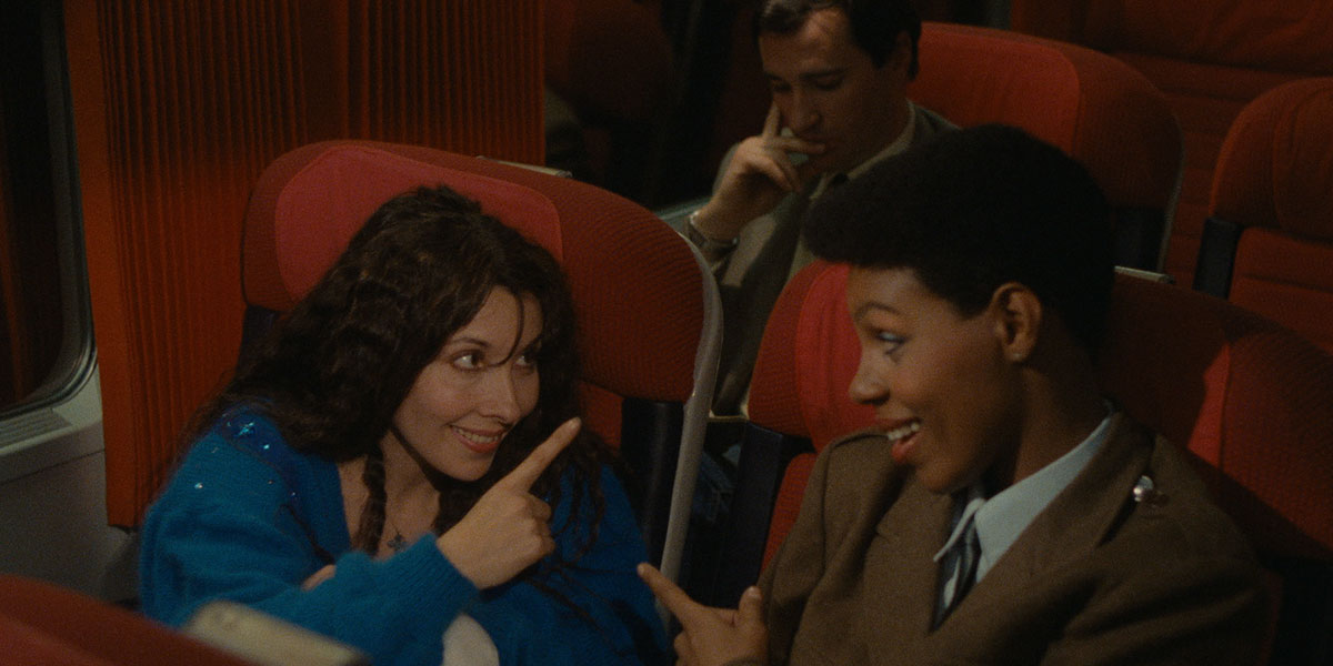 A still from Maine-Océan Express (1986) where a woman in a blue dress is pointing to a woman in a brown suit who is also pointing back. They are sitting in red seats on a train