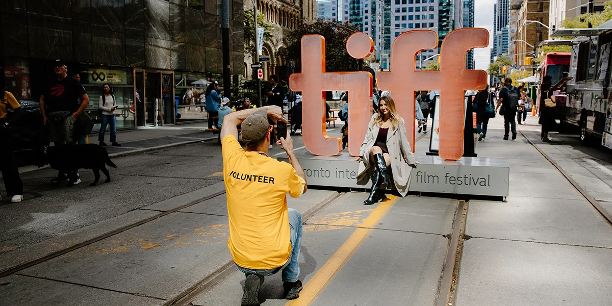 Volunteer in yellow shirt photographs TIFF sign at Toronto International Film Festival on busy city street