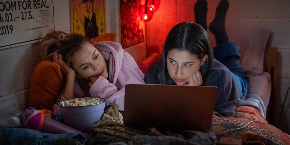 Two friends lounging together on a bed in dim red lighting, sharing popcorn while watching something on a laptop