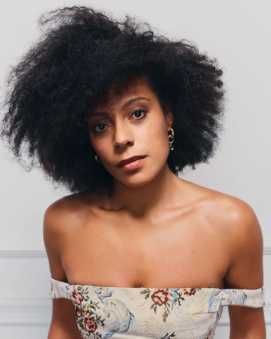 Woman with natural afro hair, gold earrings, wearing an off-shoulder floral top, neutral expression.