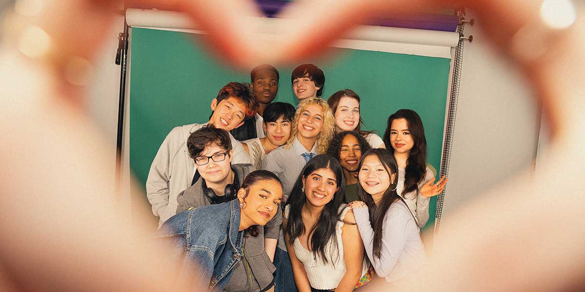 Diverse group of students smiling together for a photo against a green backdrop, with blurred edges of hands forming a heart framing the shot