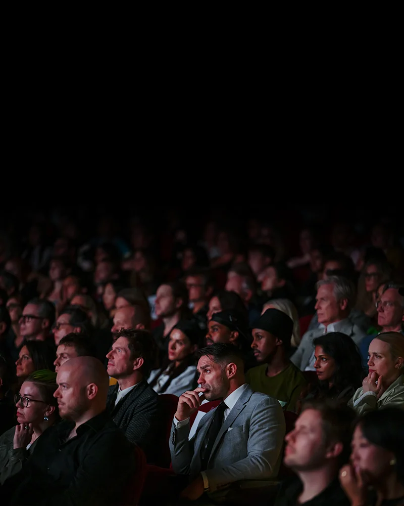 A large audience seated in a dimly lit venue, attentively watching a presentation or performance.