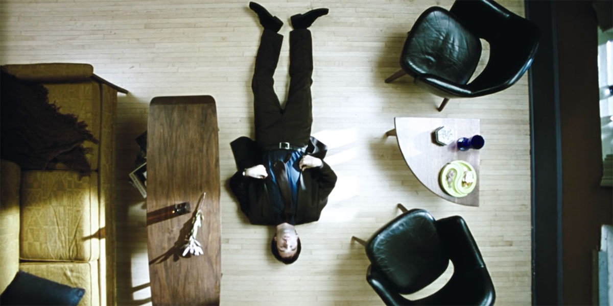 Looking down onto a man in a suit lying down on the living room floor surrounded by a coffee table, a tan couch, and some black chairs