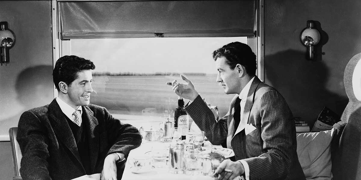 Two men in suits converse over a dining table on a train, black and white vintage photo.
