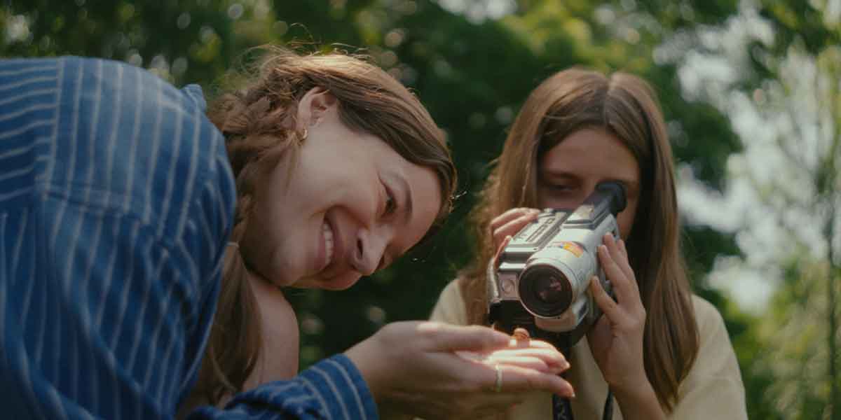 Two people smiling while filming with a handheld camcorder outdoors on a sunny day