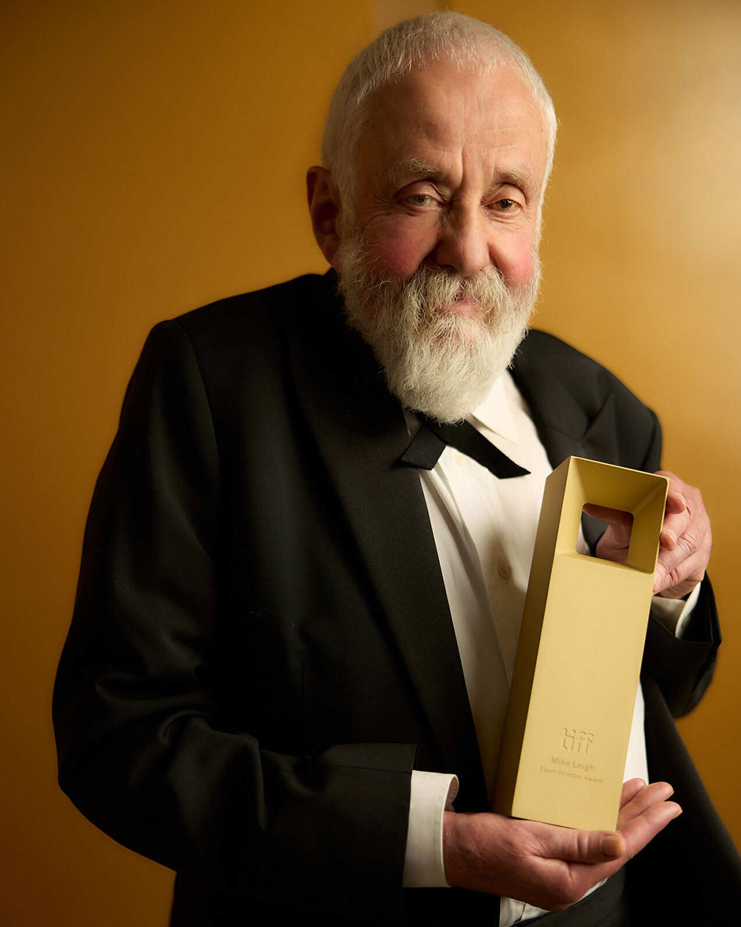 Portrait of Mike Leigh in a black suit sitting in a chair and holding his award