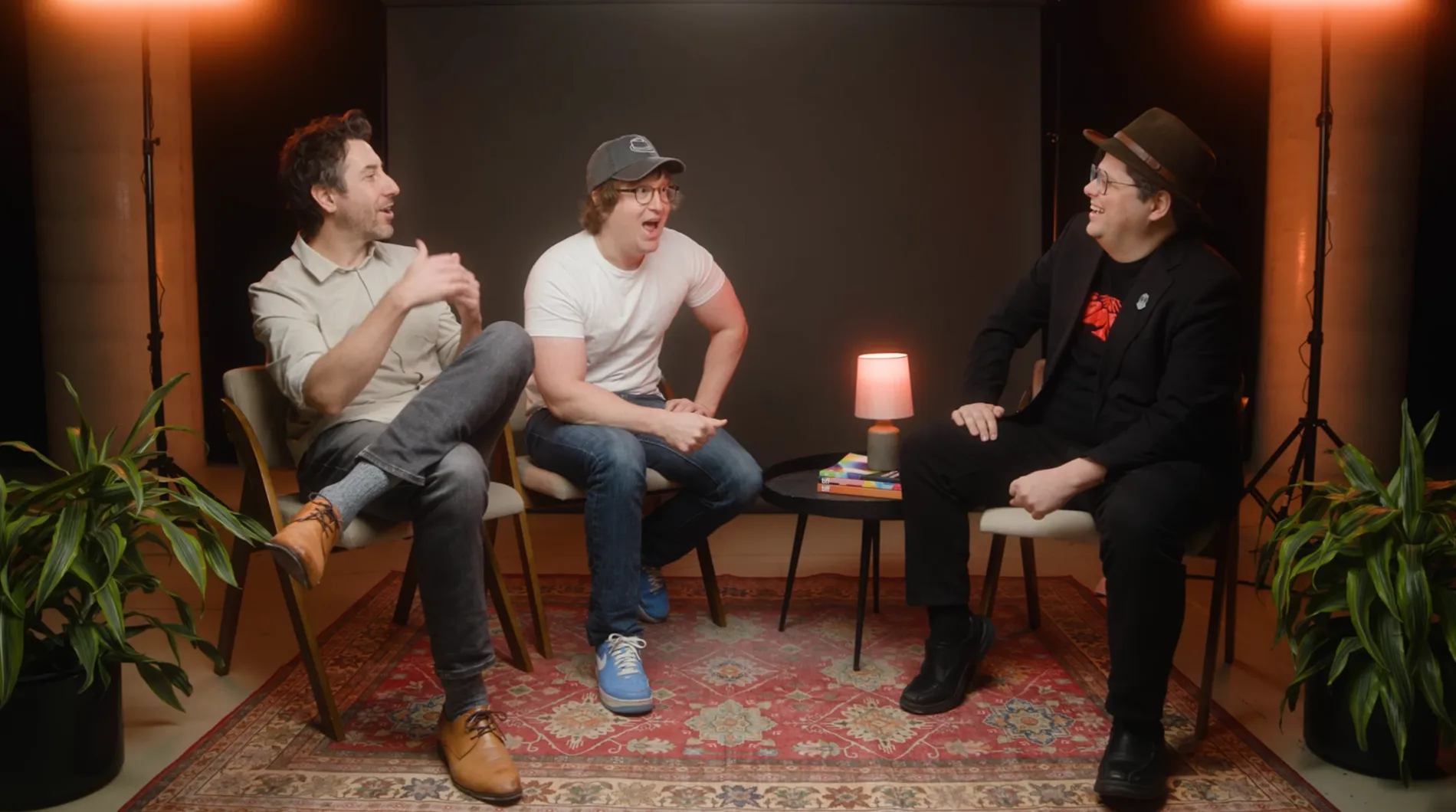 Three men sit in chairs on a studio set with bright lights, laughing and conversing together.