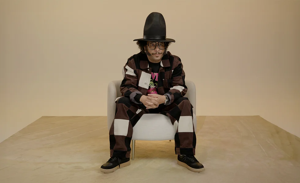 A man in a checkered brown outfit and a tall black hat sits in a white chair against a beige background.
