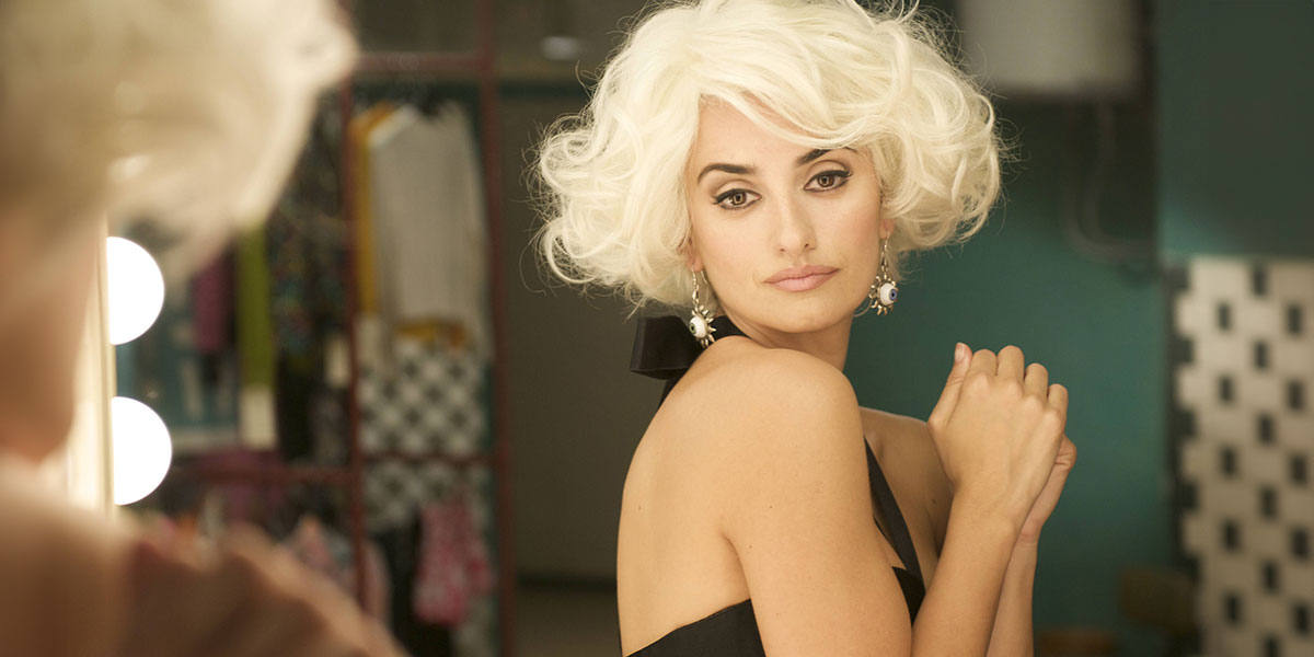 A women in a blonde wig and black dress staring at herself in the mirror inside a dressing room